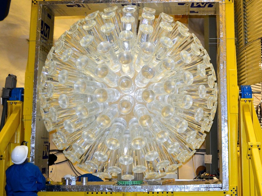 The acrylic vessel of DEAP-3600: a large acrylic sphere with 50 cm long acrylic light guides coming out of it. Someone in a blue cleanroom suit is looking up at the detector