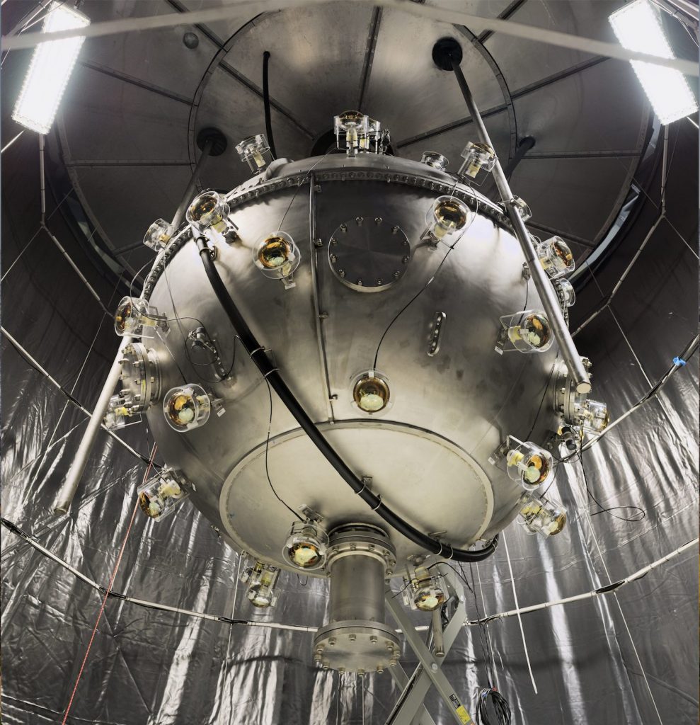 DEAP-3600 enclosed in a spherical stainless steel shell with muon veto PMTs mounted on the outside, looking into the not-yet-filled water tank, seen in the background with a black liner and scaffolding during construction